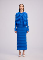 A woman faces forward in the ISSEY MIYAKE SPRING PLEATS dress—a vibrant blue, long-sleeved, floor-length piece with handmade pleats and a ribbed texture—paired with white open-toe heels against a plain light background.