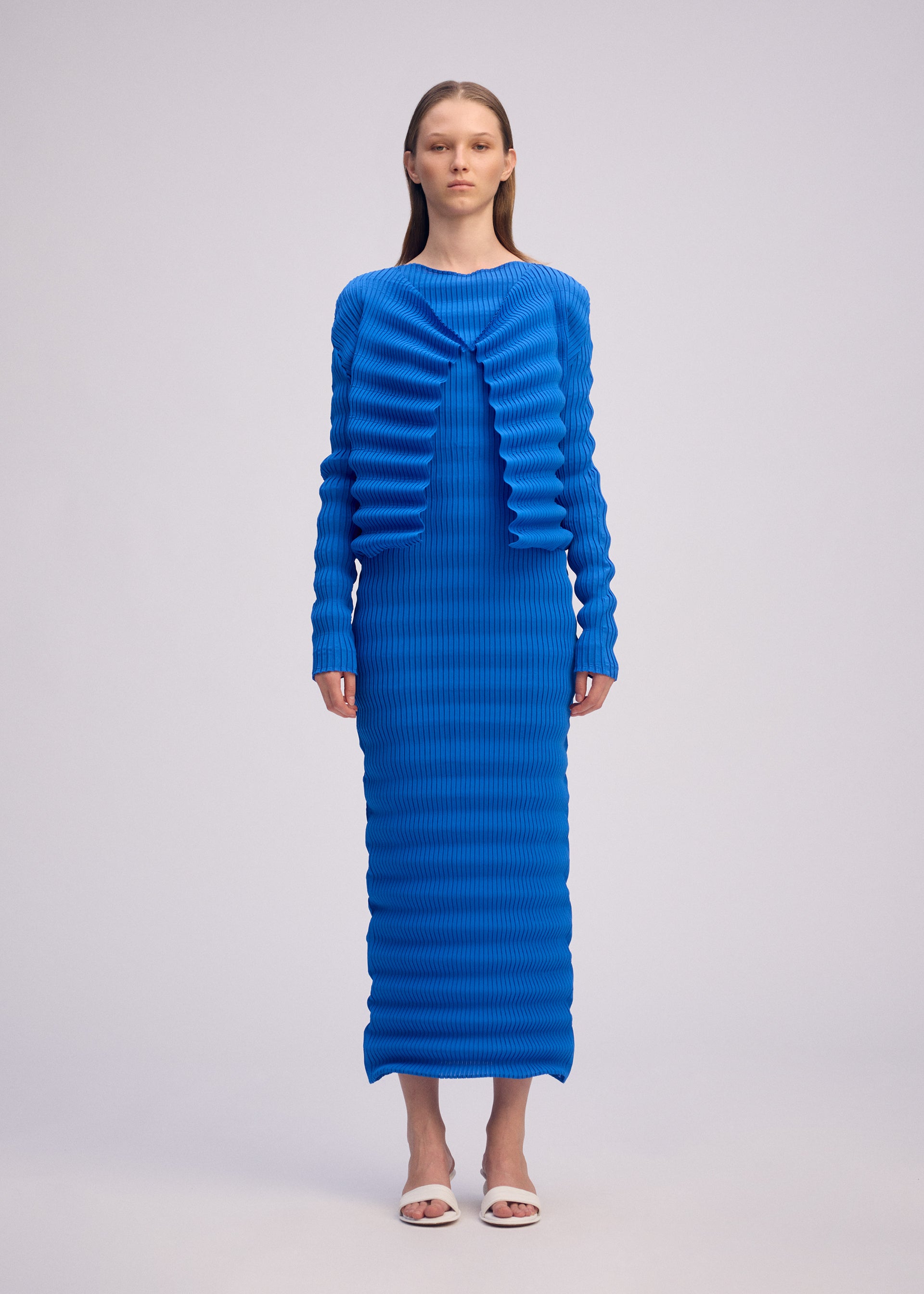 Zoomed view A woman faces forward in the ISSEY MIYAKE SPRING PLEATS dress—a vibrant blue, long-sleeved, floor-length piece with handmade pleats and a ribbed texture—paired with white open-toe heels against a plain light background.