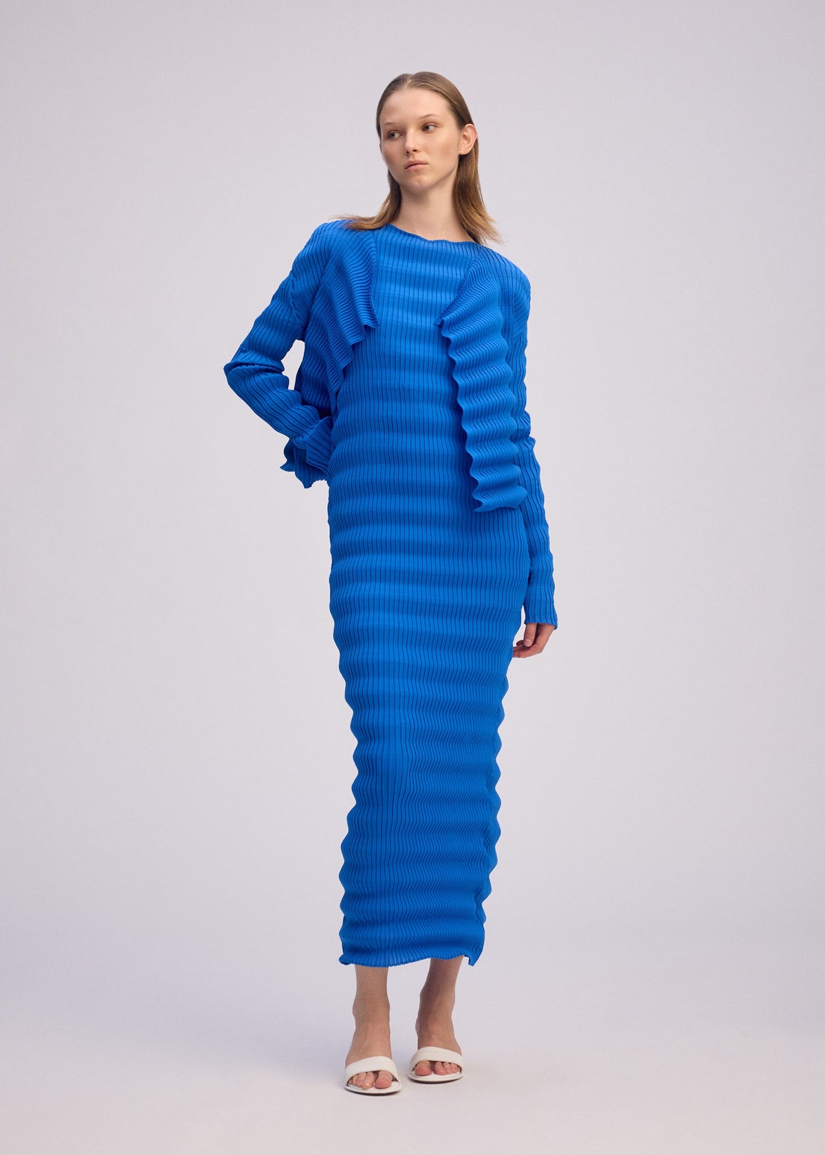 A woman poses against a plain background in the ISSEY MIYAKE SPRING PLEATS dress—a long, bright blue piece with handmade pleats and wavy textures. She styles it with white open-toe heels, long sleeves, and straight light brown hair. view 5 of 6