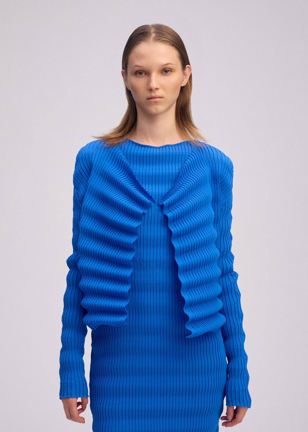 A person with straight, light brown hair models the ISSEY MIYAKE SPRING PLEATS—a bright blue, long-sleeve ribbed dress and matching cardigan featuring handmade pleats and wavy details—against a plain light background. view 6 of 6