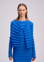 A person with straight, light brown hair models the ISSEY MIYAKE SPRING PLEATS—a bright blue, long-sleeve ribbed dress and matching cardigan featuring handmade pleats and wavy details—against a plain light background.