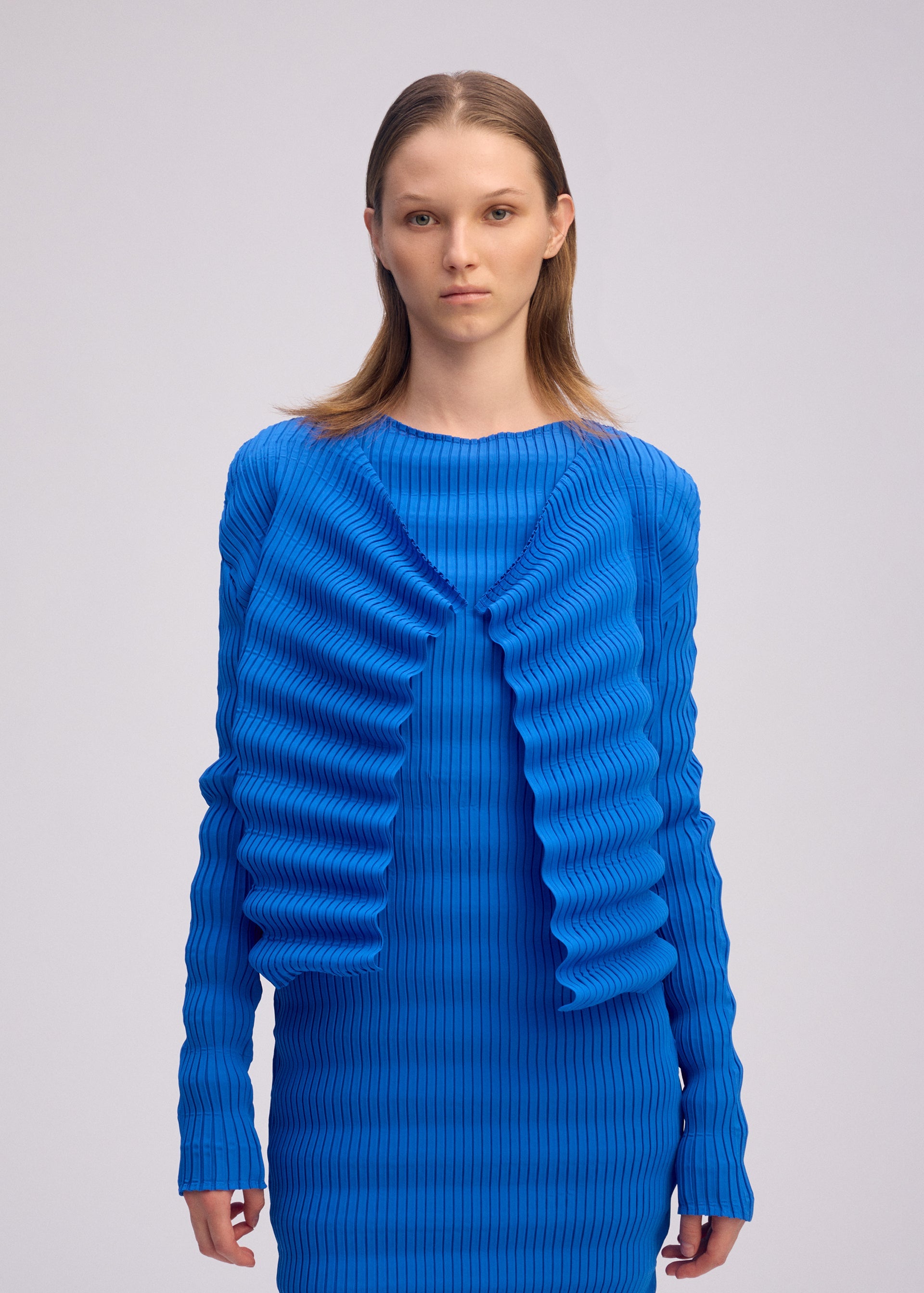 Zoomed view A person with straight, light brown hair models the ISSEY MIYAKE SPRING PLEATS—a bright blue, long-sleeve ribbed dress and matching cardigan featuring handmade pleats and wavy details—against a plain light background.