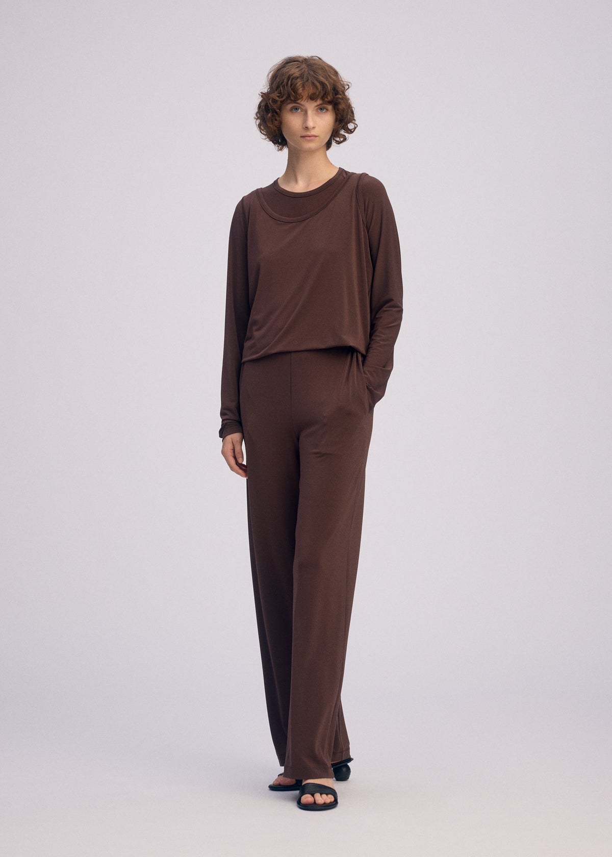 A person with short curly hair stands against a plain light background, wearing the ISSEY MIYAKE DRAPE JERSEY-66 top and wide-leg pants, paired with black open-toe sandals. One hand in their pocket adds to the flowy feel of the look. view 6 of 8