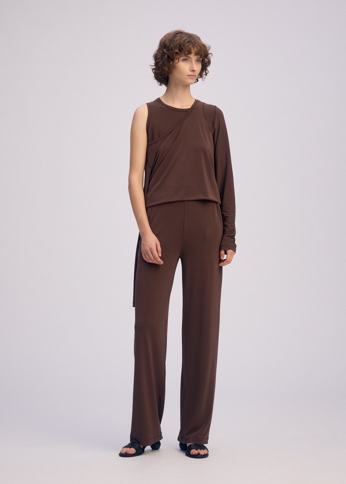 A person with short curly hair stands against a plain background, wearing the ISSEY MIYAKE DRAPE JERSEY-66 asymmetrical long-sleeve top and matching straight pants, styled with black sandals. view 7 of 8
