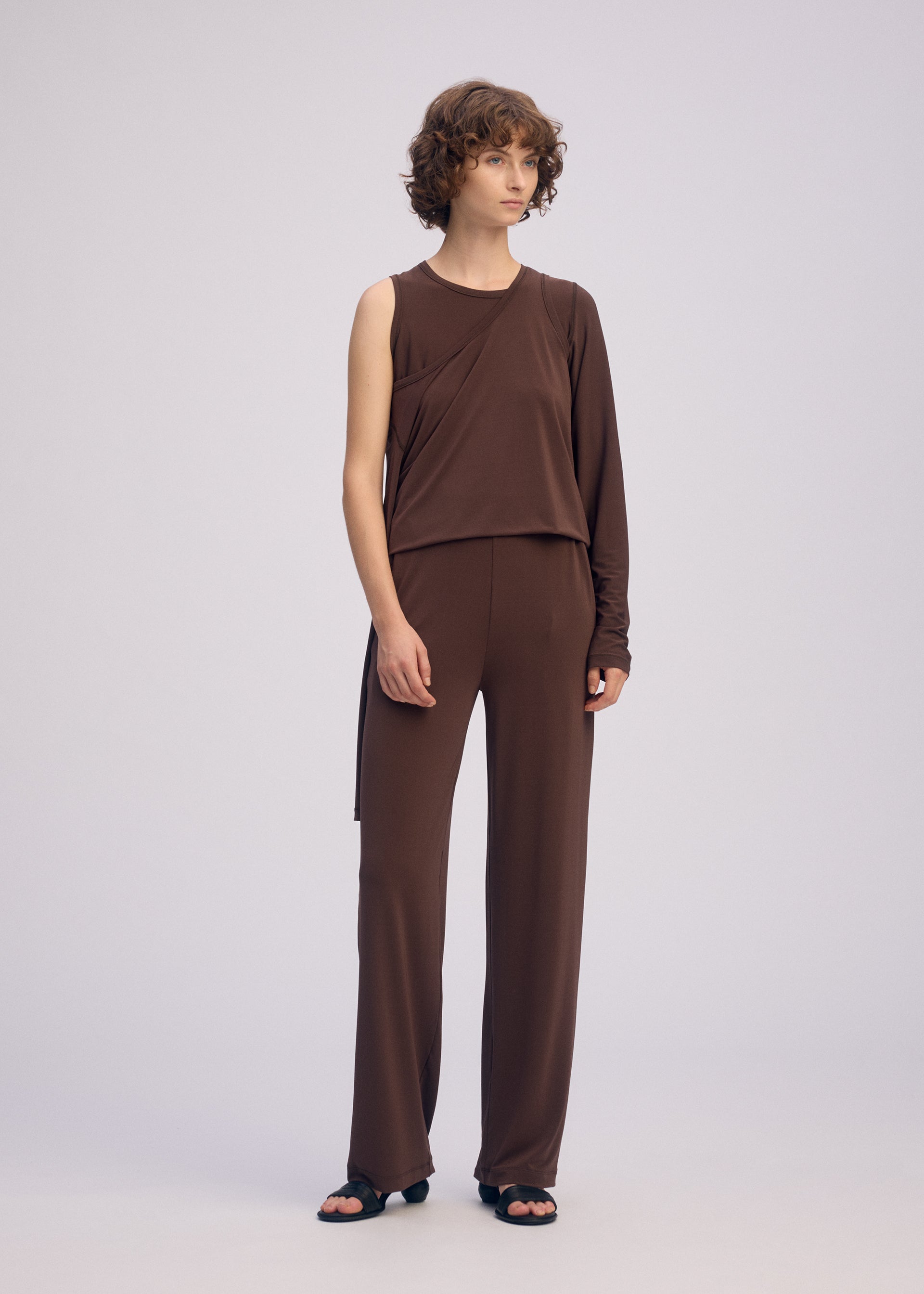 Zoomed view A person with short curly hair stands against a plain background, wearing the ISSEY MIYAKE DRAPE JERSEY-66 asymmetrical long-sleeve top and matching straight pants, styled with black sandals.