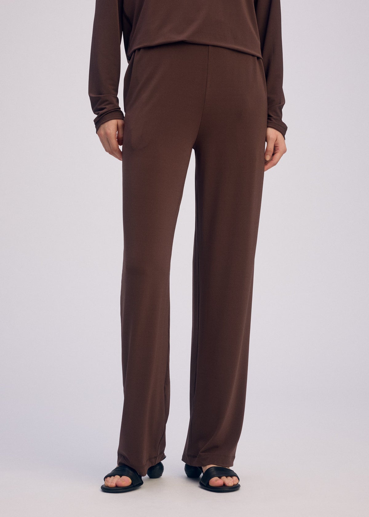 The lower half of a person is shown wearing ISSEY MIYAKE's DRAPE JERSEY-66 loose brown straight pants and matching long-sleeve top, set against a plain light background, with black open-toe sandals emphasizing the flowy silhouette. view 8 of 8