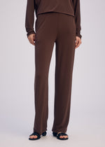 The lower half of a person is shown wearing ISSEY MIYAKE's DRAPE JERSEY-66 loose brown straight pants and matching long-sleeve top, set against a plain light background, with black open-toe sandals emphasizing the flowy silhouette.