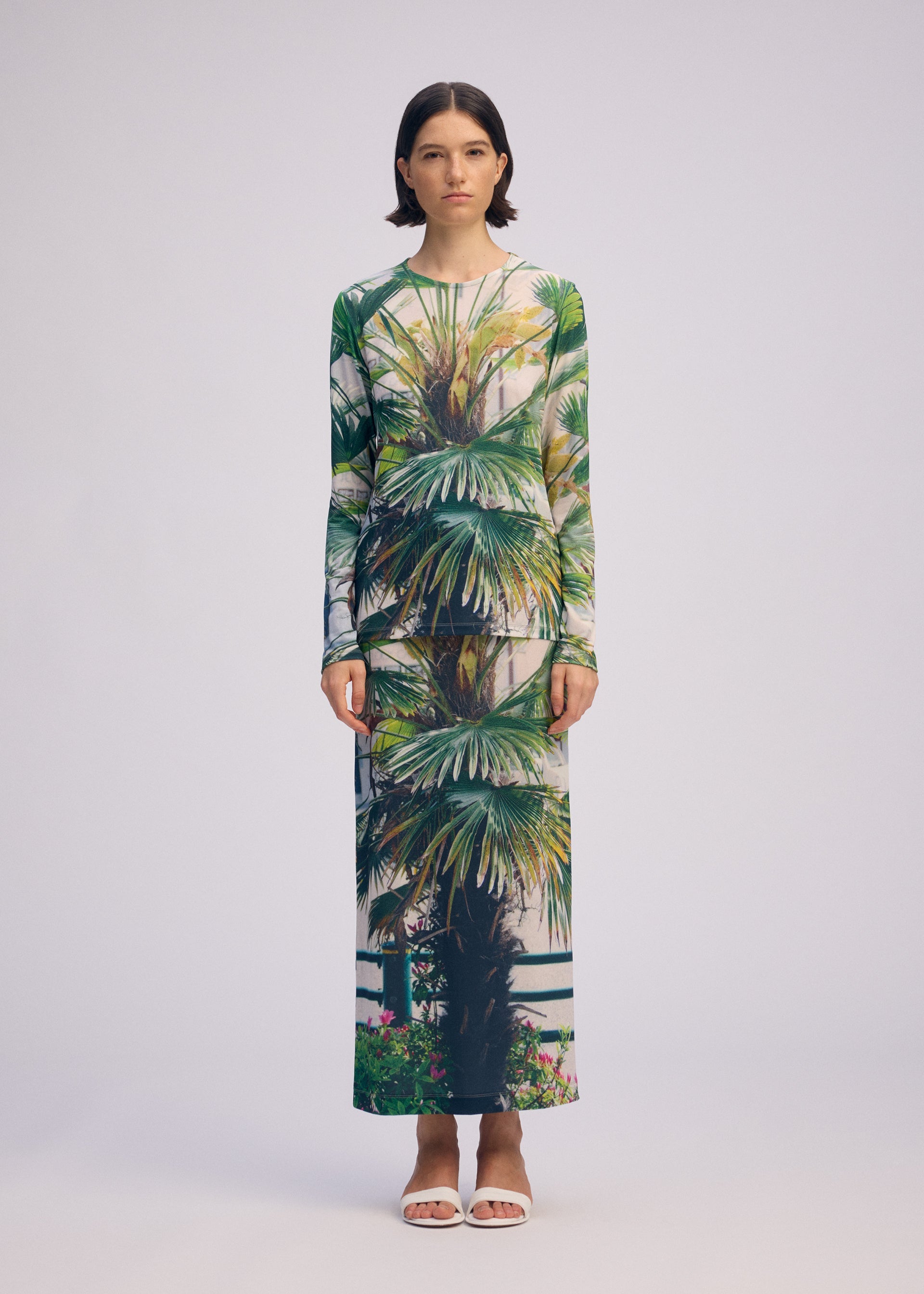 Zoomed view A woman with straight dark hair wears the ISSEY MIYAKE URBAN JUNGLE JERSEY—a vivid palm print long-sleeved top and matching skirt—against a plain background, paired with white open-toe sandals.