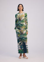 A woman with short dark hair wears the ISSEY MIYAKE URBAN JUNGLE JERSEY, a long-sleeved, ankle-length dress with a palm plant pattern and lush greenery, paired with white sandals against a plain background.