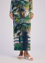 Wearing the ISSEY MIYAKE URBAN JUNGLE JERSEY set with vibrant palm patterns and white open-toe heeled sandals, a person stands against a plain light background.