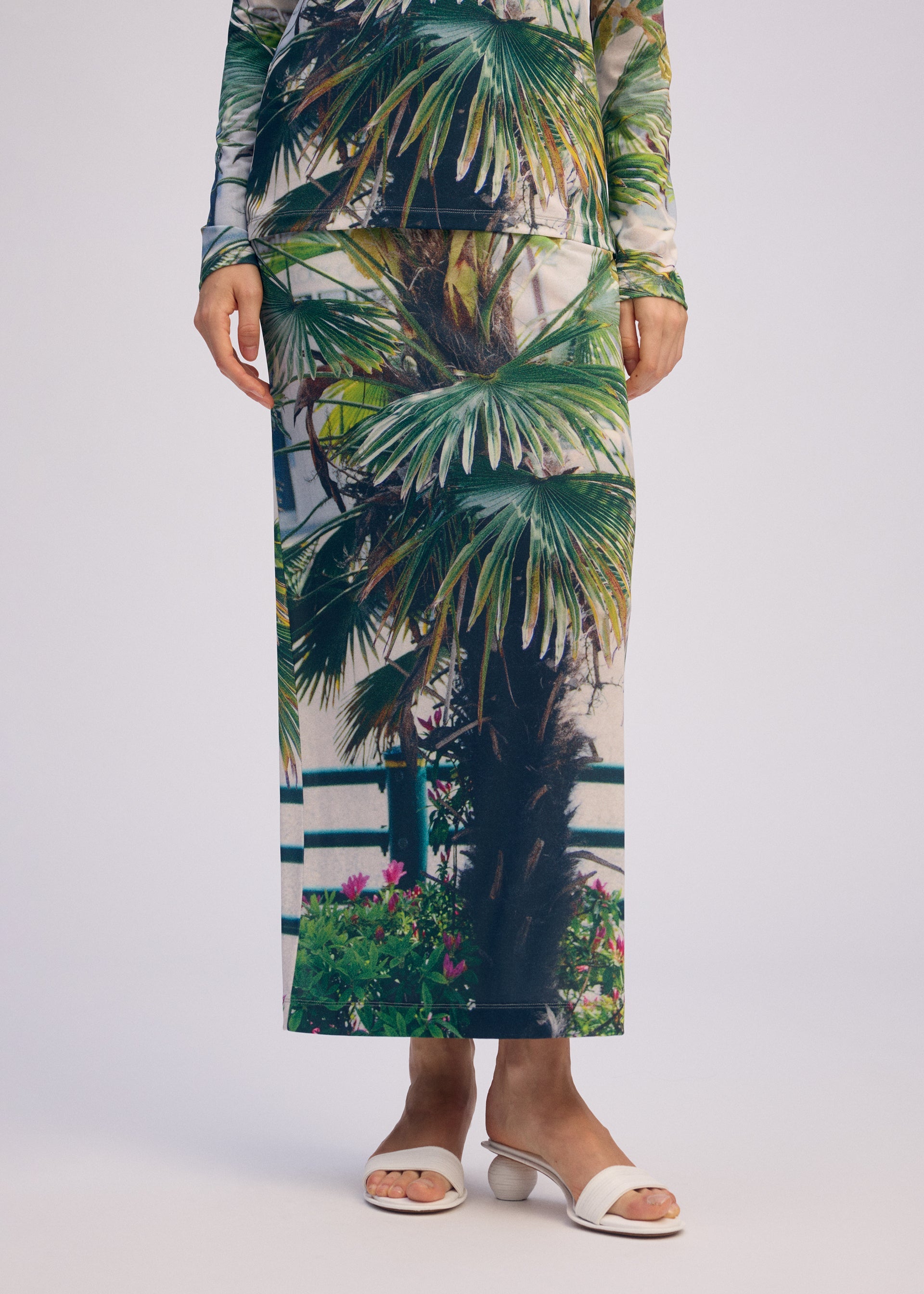 Zoomed view Wearing the ISSEY MIYAKE URBAN JUNGLE JERSEY set with vibrant palm patterns and white open-toe heeled sandals, a person stands against a plain light background.