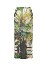 The URBAN JUNGLE JERSEY by ISSEY MIYAKE is a fitted long skirt showcasing a lifelike palm plant design with a brown trunk, light background, and accents of railing and foliage along the hem.