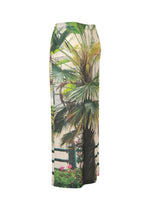 The ISSEY MIYAKE URBAN JUNGLE JERSEY is a long skirt with a bold palm plant print featuring green foliage, floral accents, and a fence motif, shown against a white background.