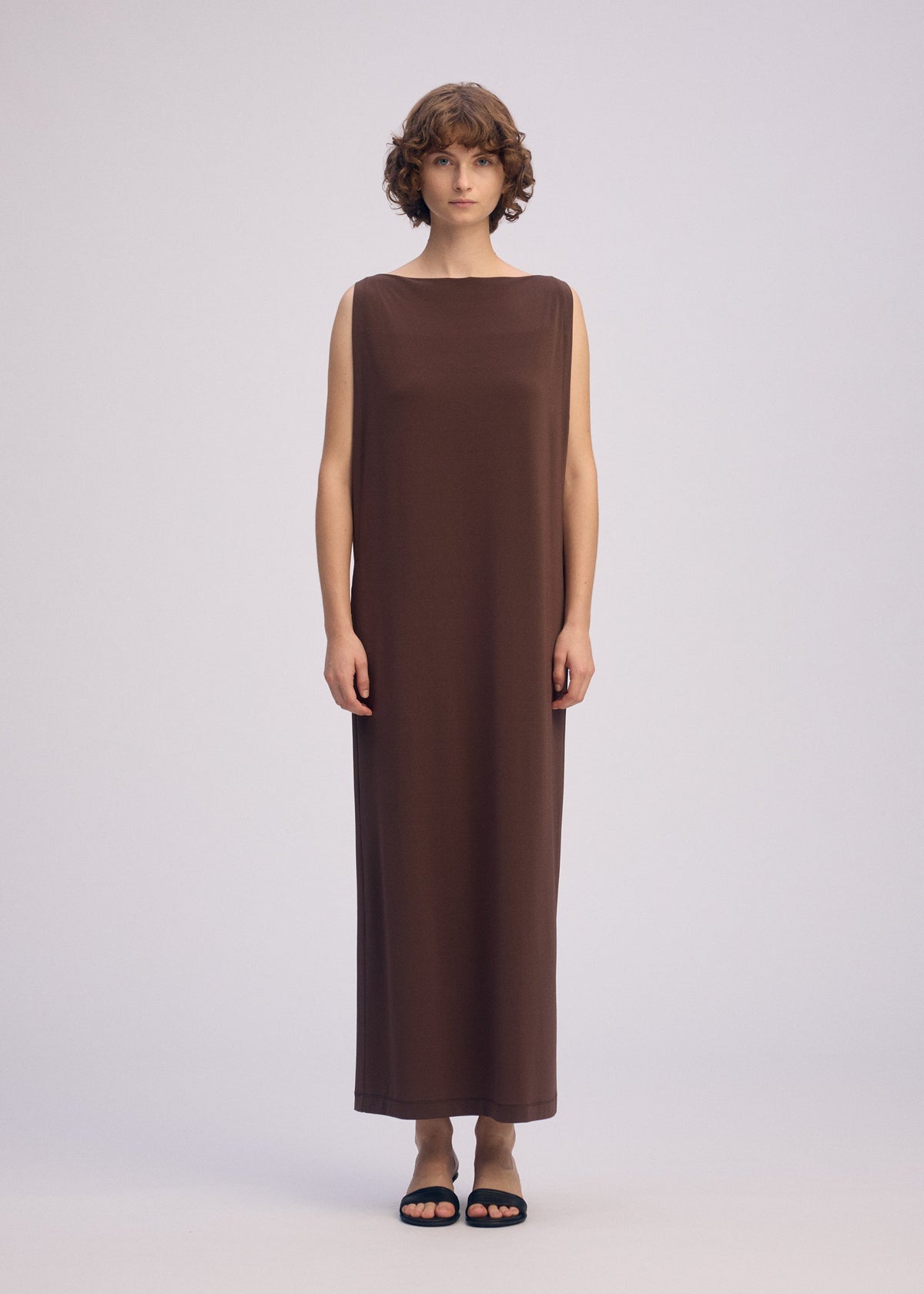 A person with short, curly hair wears the ISSEY MIYAKE DRAPE JERSEY-66, a sleeveless flowy dress in brown, paired with black sandals, standing against a plain light background. view 6 of 7