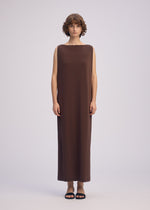 A person with short, curly hair wears the ISSEY MIYAKE DRAPE JERSEY-66, a sleeveless flowy dress in brown, paired with black sandals, standing against a plain light background.