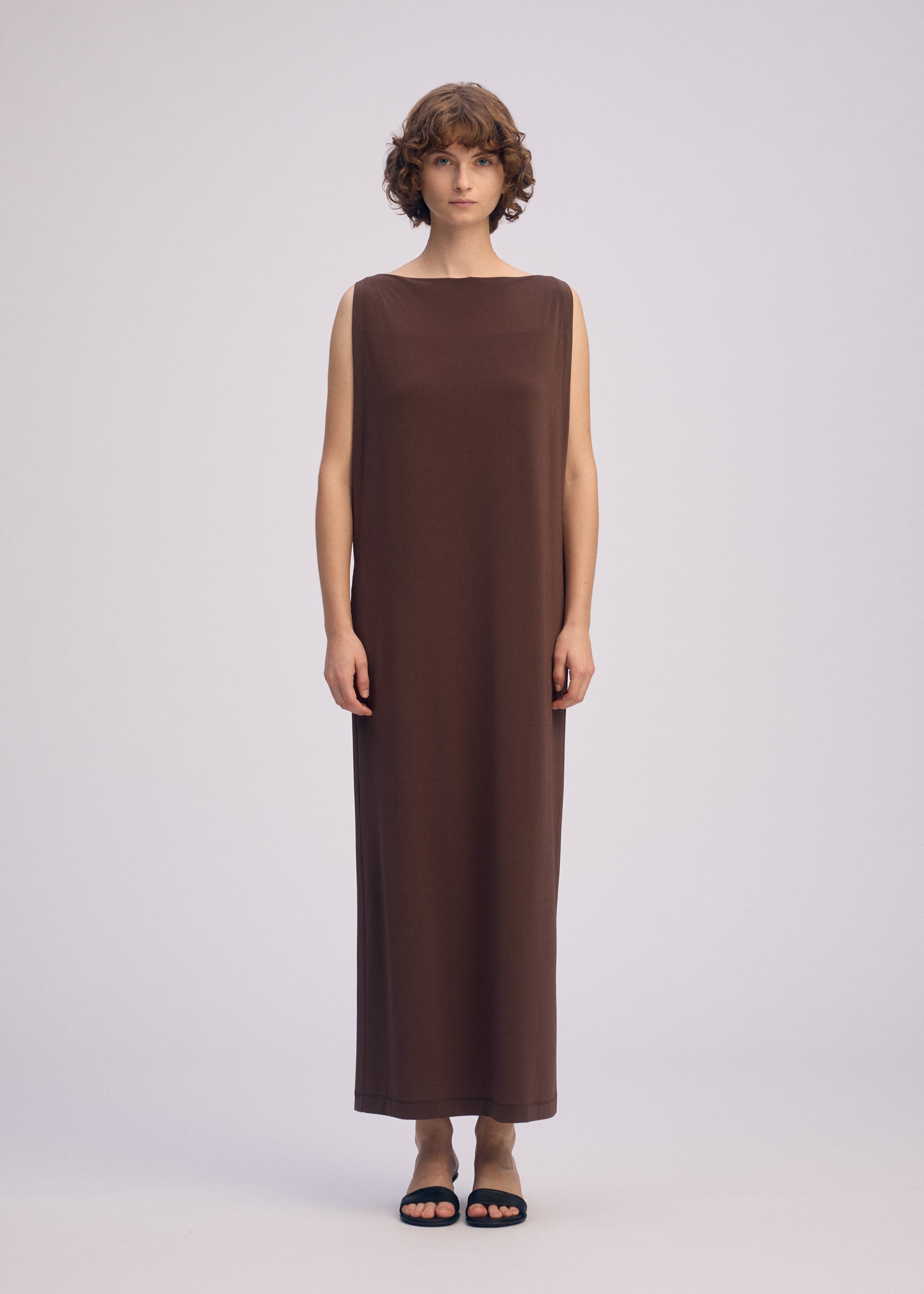 Zoomed view A person with short, curly hair wears the ISSEY MIYAKE DRAPE JERSEY-66, a sleeveless flowy dress in brown, paired with black sandals, standing against a plain light background.