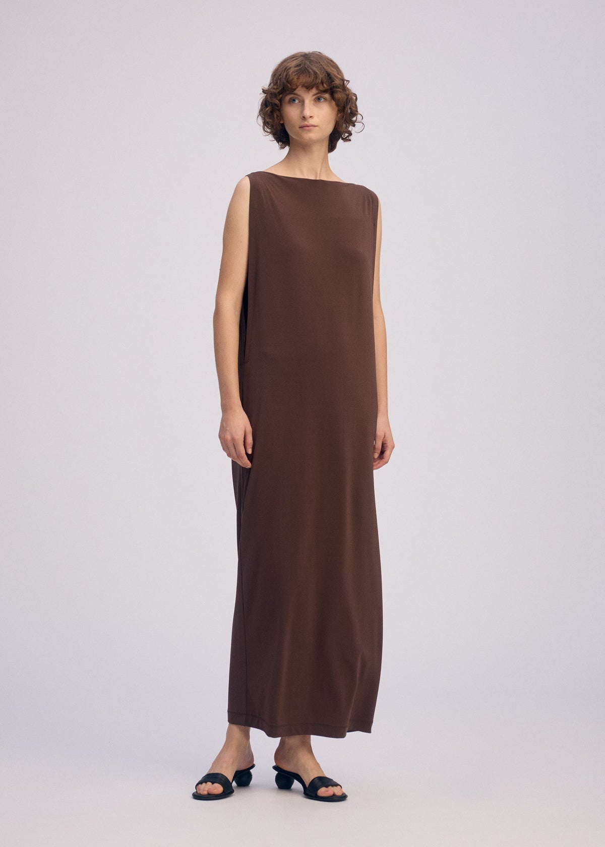 A person with short curly hair wears the DRAPE JERSEY-66 sleeveless dress by ISSEY MIYAKE and black open-toe heeled sandals, standing against a plain light background. view 7 of 7