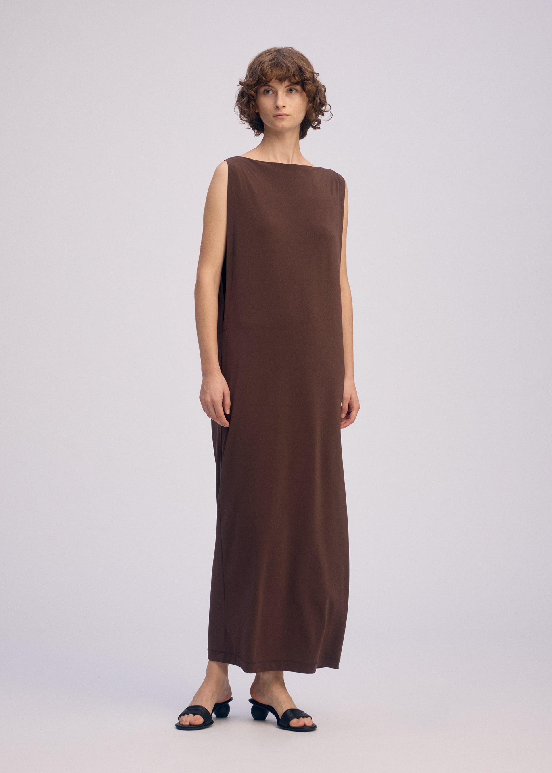 Zoomed view A person with short curly hair wears the DRAPE JERSEY-66 sleeveless dress by ISSEY MIYAKE and black open-toe heeled sandals, standing against a plain light background.