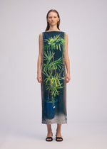 A woman poses against a plain background in the URBAN JUNGLE JERSEY dress by ISSEY MIYAKE, a sleeveless, floor-length piece crafted from soft jersey and printed with jungle greenery in a blue pot. She faces forward with a neutral expression.