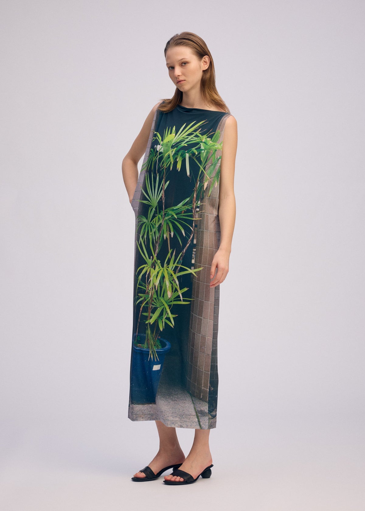A woman wears the ISSEY MIYAKE URBAN JUNGLE JERSEY, a sleeveless dress in soft jersey with a jungle greenery print and potted plant motif, standing before a plain light backdrop. view 6 of 6