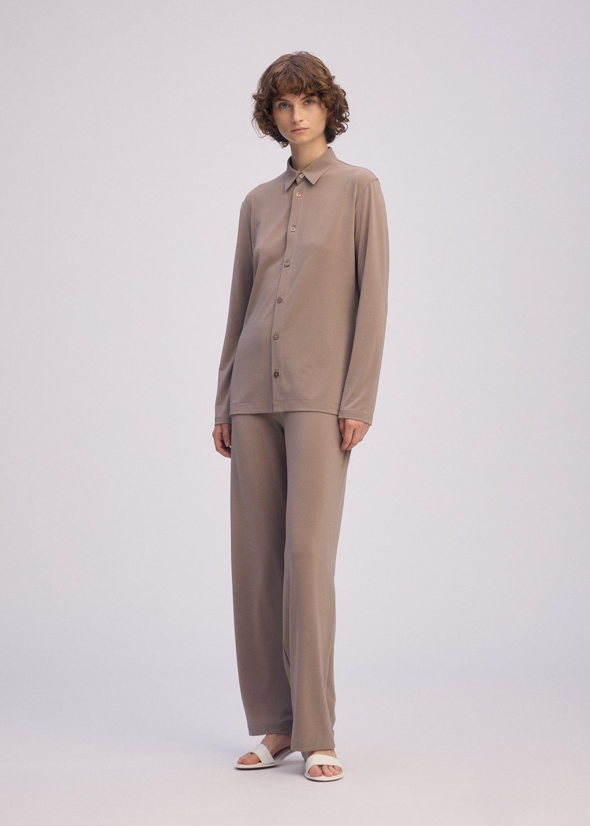 A person with short curly hair wears the taupe DRAPE JERSEY-66 flowy shirt and matching wide-leg pants by ISSEY MIYAKE, paired with white open-toe sandals, standing against a plain light background. view 6 of 7