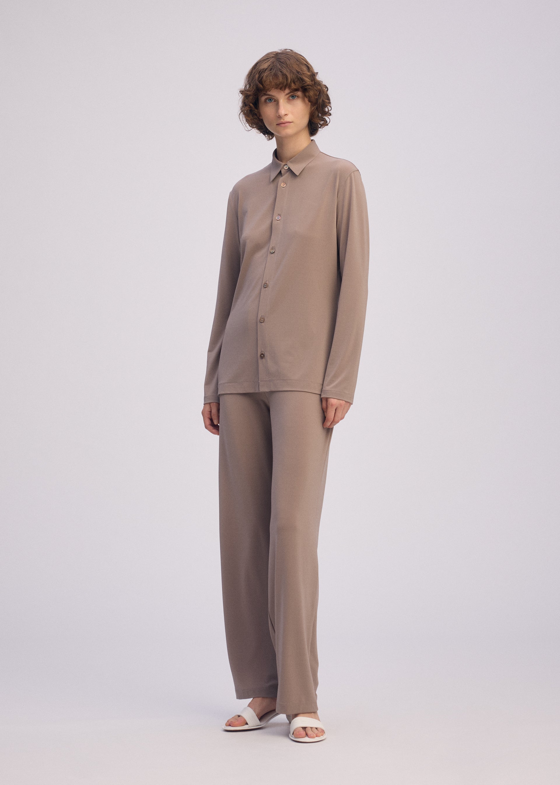 Zoomed view A person with short curly hair wears the taupe DRAPE JERSEY-66 flowy shirt and matching wide-leg pants by ISSEY MIYAKE, paired with white open-toe sandals, standing against a plain light background.