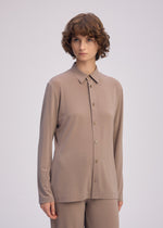 A person with short, curly brown hair wears the loose-fitting DRAPE JERSEY-66 taupe shirt and matching pants by ISSEY MIYAKE, standing against a plain light background.