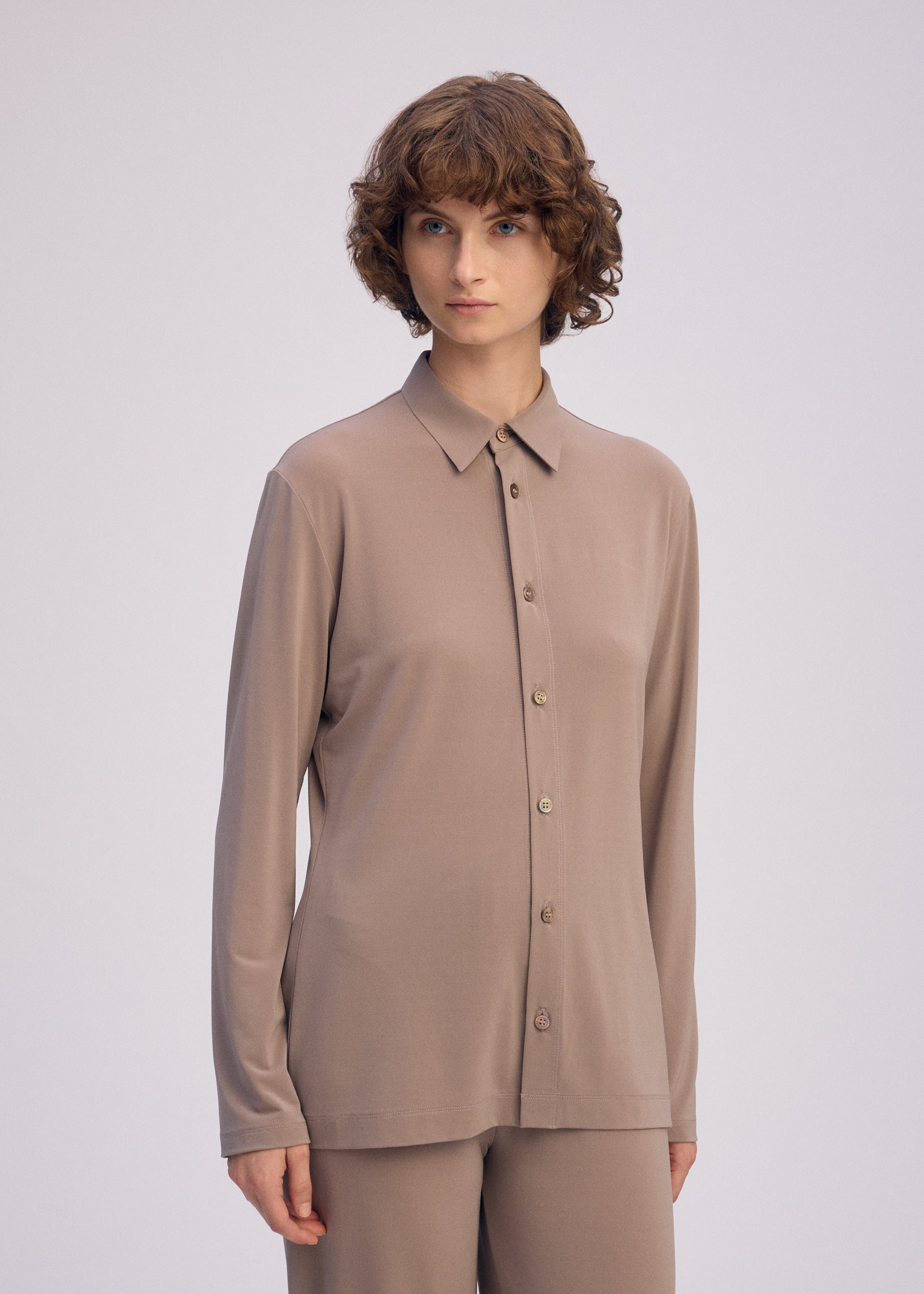 Zoomed view A person with short, curly brown hair wears the loose-fitting DRAPE JERSEY-66 taupe shirt and matching pants by ISSEY MIYAKE, standing against a plain light background.
