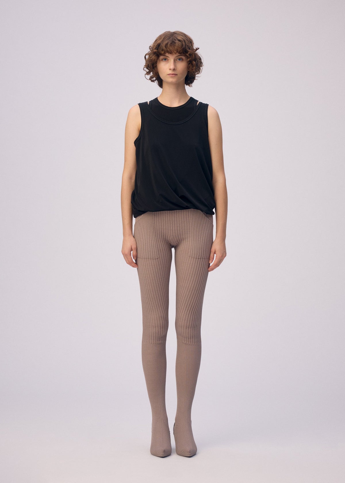 A person with short curly hair stands against a plain background, wearing the ISSEY MIYAKE DRAPE JERSEY-66 sleeveless top and taupe ribbed tights. They face forward with a neutral expression and arms relaxed at their sides. view 5 of 8