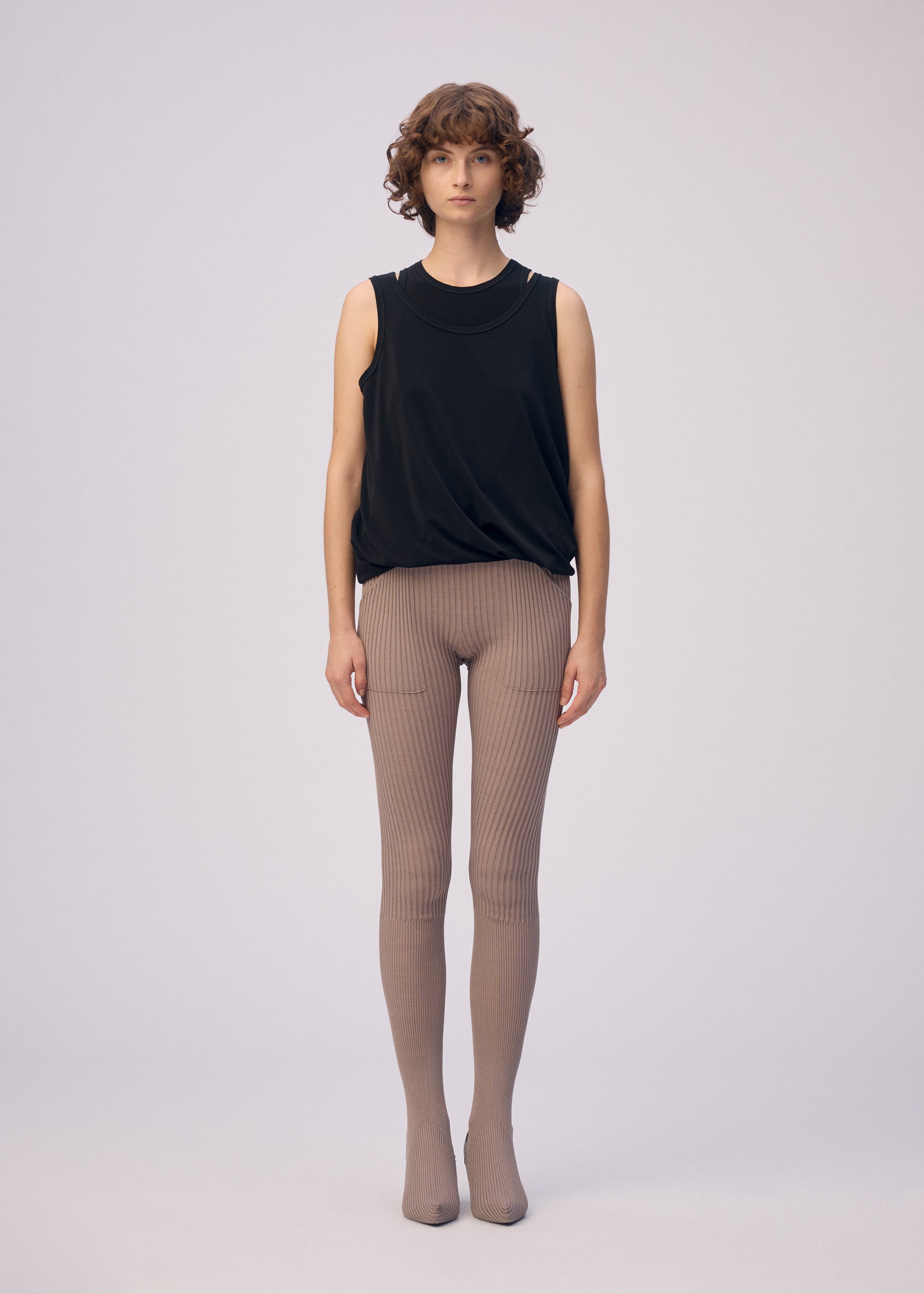 Zoomed view A person with short curly hair stands against a plain background, wearing the ISSEY MIYAKE DRAPE JERSEY-66 sleeveless top and taupe ribbed tights. They face forward with a neutral expression and arms relaxed at their sides.
