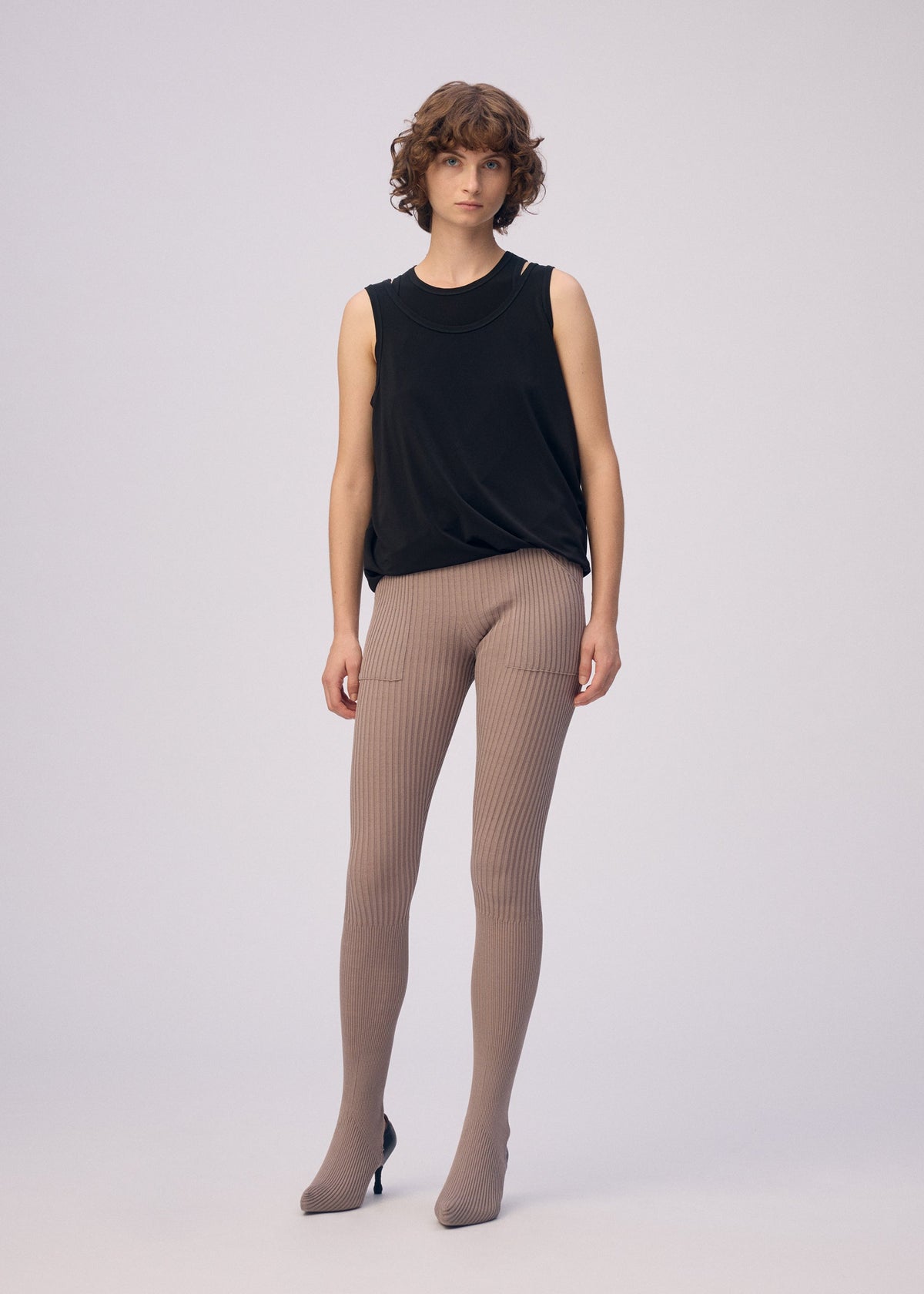 A person with short, wavy brown hair stands against a plain light background, wearing the ISSEY MIYAKE DRAPE JERSEY-66 sleeveless top and beige ribbed leggings with pointed-toe shoes, highlighting the outfit's modern design. view 6 of 8