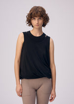 A person with short curly brown hair wears the ISSEY MIYAKE DRAPE JERSEY-66 sleeveless black top and beige ribbed pants with a subtle layered design, standing against a plain light background and facing the camera neutrally.