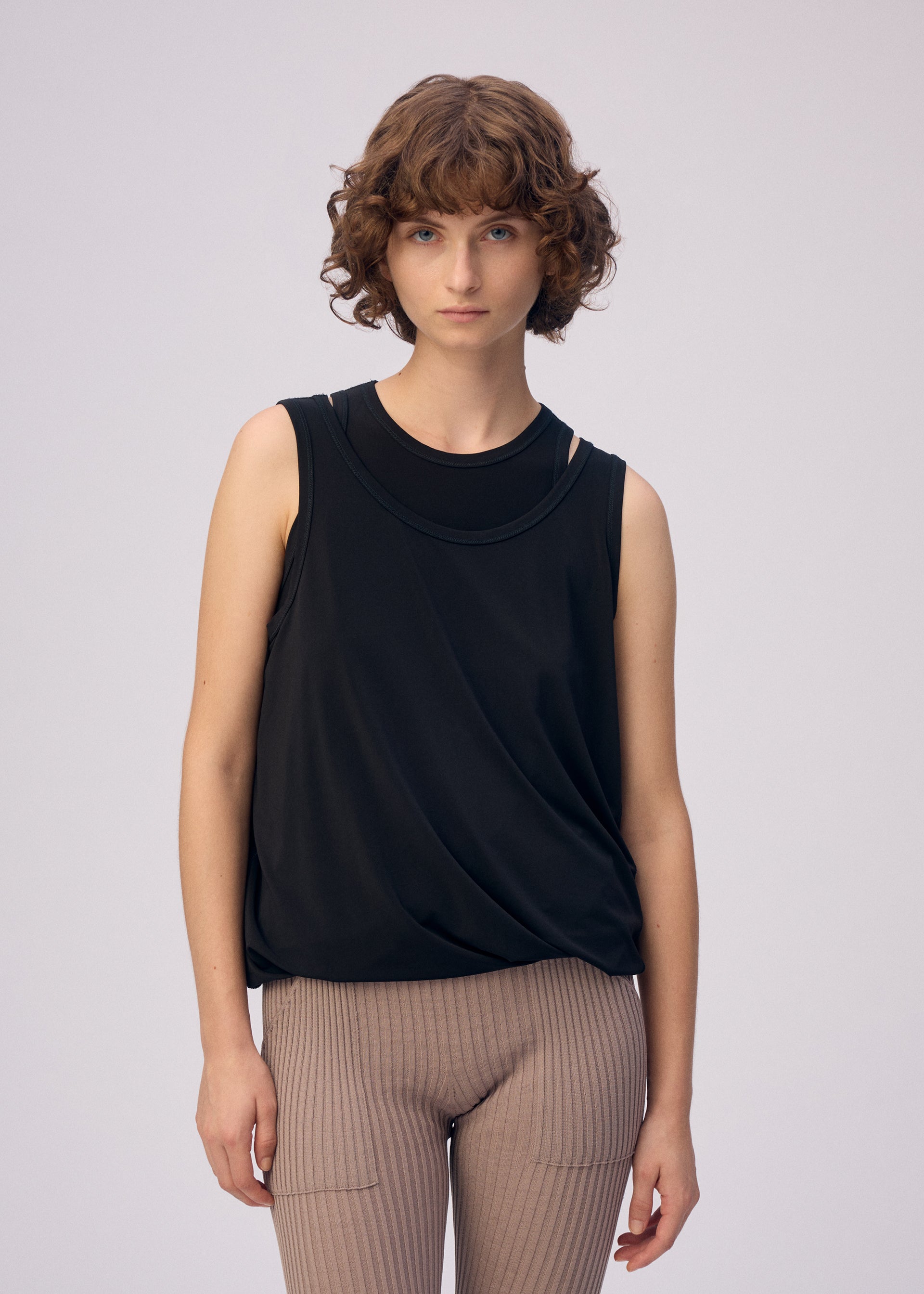 Zoomed view A person with short curly brown hair wears the ISSEY MIYAKE DRAPE JERSEY-66 sleeveless black top and beige ribbed pants with a subtle layered design, standing against a plain light background and facing the camera neutrally.