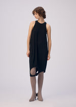 A woman with short curly hair models the ISSEY MIYAKE DRAPE JERSEY-66 sleeveless top layered over a black dress with an asymmetrical hem, taupe ribbed tights, and black pointed heels, standing against a plain light backdrop, looking left.