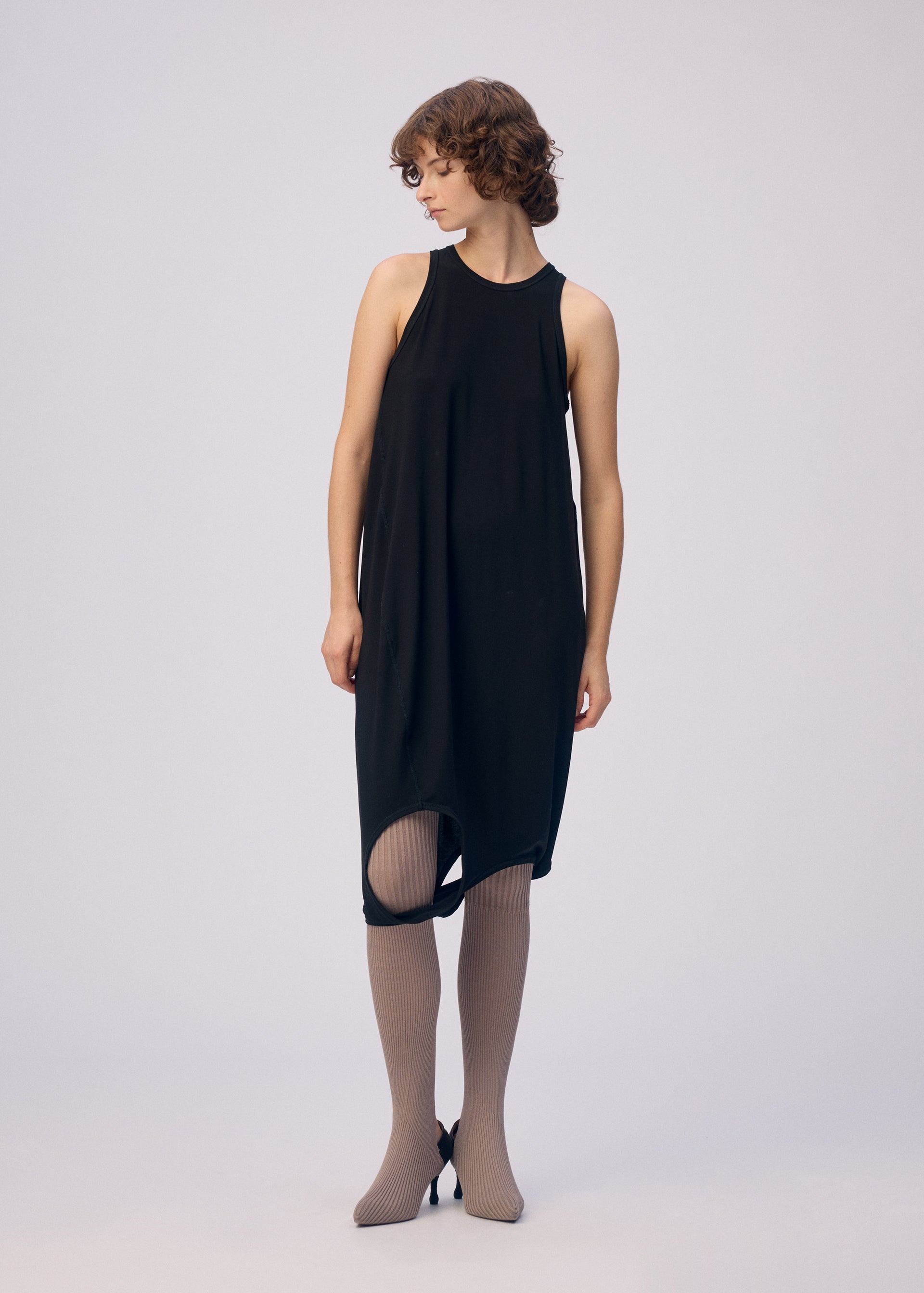 Zoomed view A woman with short curly hair models the ISSEY MIYAKE DRAPE JERSEY-66 sleeveless top layered over a black dress with an asymmetrical hem, taupe ribbed tights, and black pointed heels, standing against a plain light backdrop, looking left.