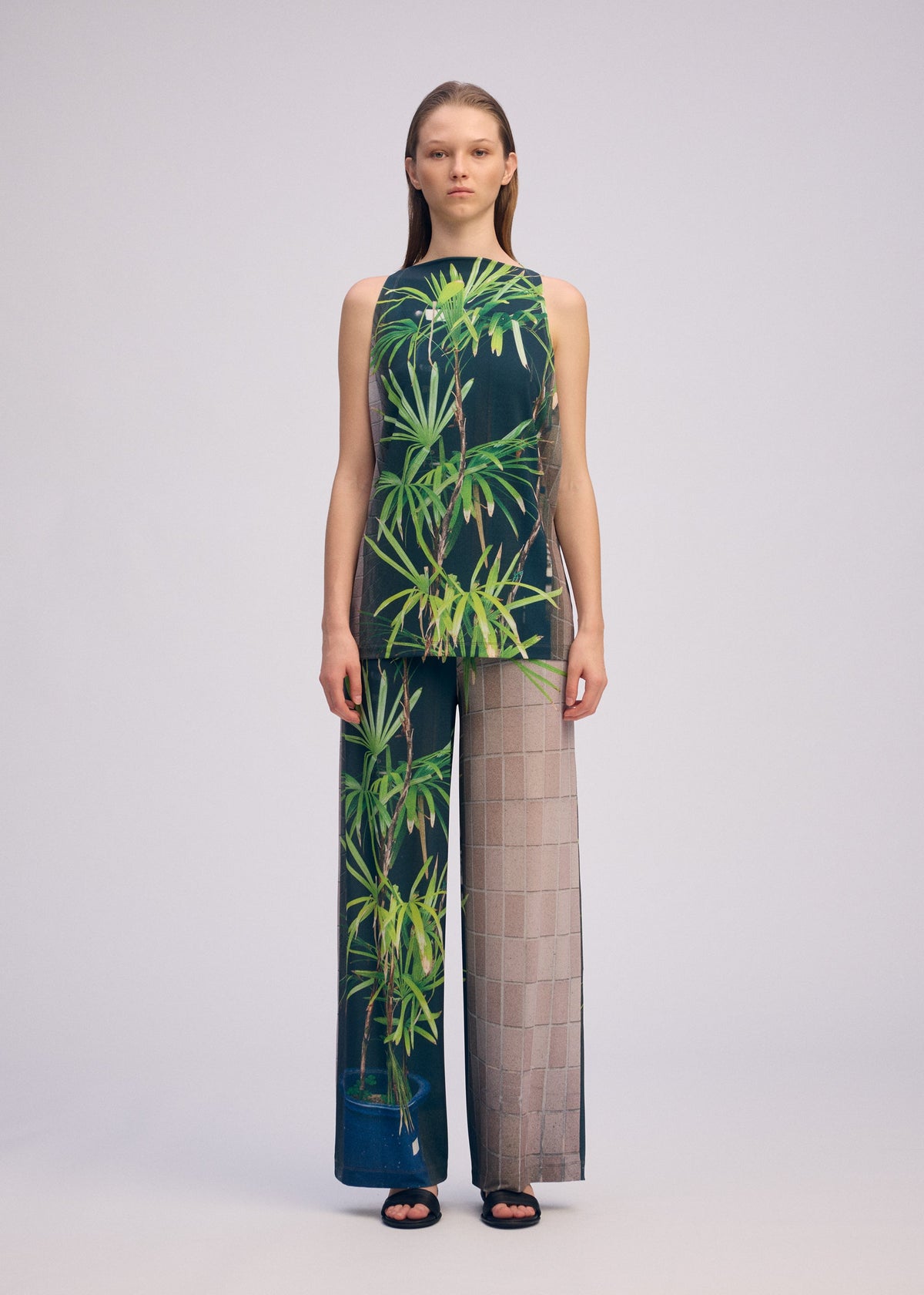 A woman faces forward in the ISSEY MIYAKE URBAN JUNGLE JERSEY, a sleeveless top and wide-leg pants set featuring green leafy jungle print on one side and a sheer beige grid pattern on the other, paired with black sandals. view 5 of 8