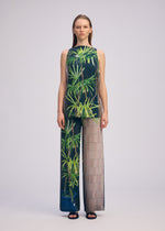 A woman faces forward in the ISSEY MIYAKE URBAN JUNGLE JERSEY, a sleeveless top and wide-leg pants set featuring green leafy jungle print on one side and a sheer beige grid pattern on the other, paired with black sandals.