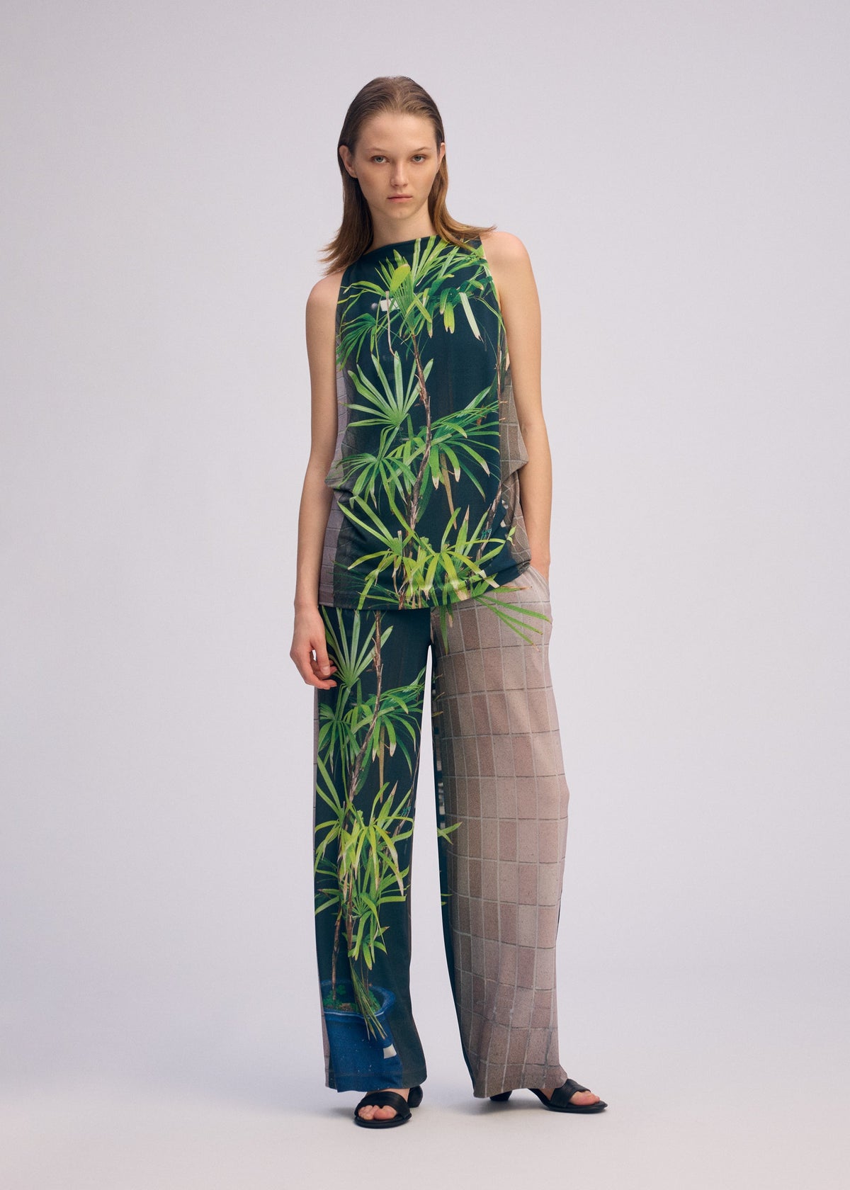 A woman with straight hair and a neutral expression wears the ISSEY MIYAKE URBAN JUNGLE JERSEY, featuring a sleeveless top and wide-leg pants with green jungle print on one side and gray checks on the other, set against a light background. view 6 of 8