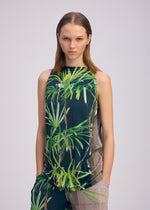A person with straight, light brown hair wears ISSEY MIYAKE's URBAN JUNGLE JERSEY sleeveless top and matching pants in a green jungle print, standing against a plain light background with hands in pockets, looking directly at the camera.