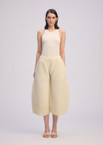 A woman stands facing forward in a sleeveless cream CORRUGATED KNIT top and wide-leg cropped pants crafted with Japanese Washi paper by ISSEY MIYAKE, paired with sandals. The background is plain and light-colored.