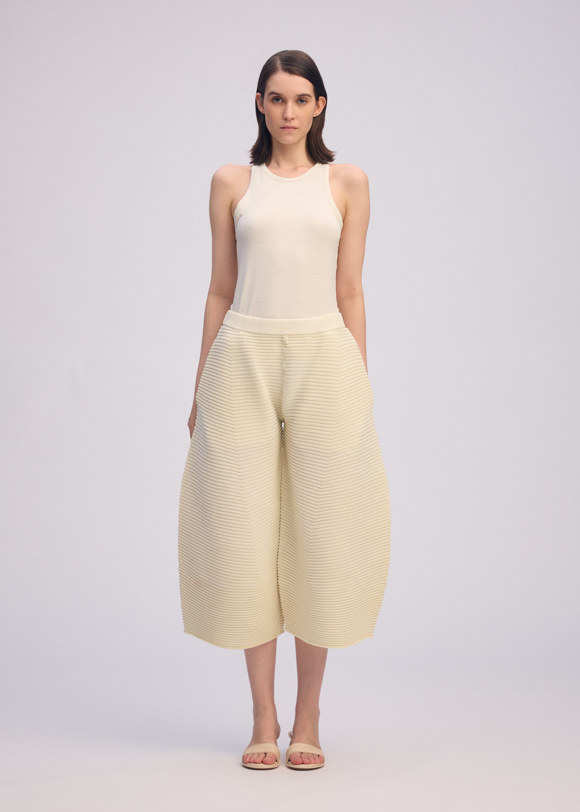 Zoomed view A woman stands facing forward in a sleeveless cream CORRUGATED KNIT top and wide-leg cropped pants crafted with Japanese Washi paper by ISSEY MIYAKE, paired with sandals. The background is plain and light-colored.