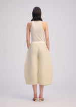 A person stands against a plain light background wearing the ISSEY MIYAKE CORRUGATED KNIT sleeveless ivory top, wide pleated culottes made from Japanese Washi paper, and beige sandals. Their hands are in their pockets and their straight hair is shoulder-length.