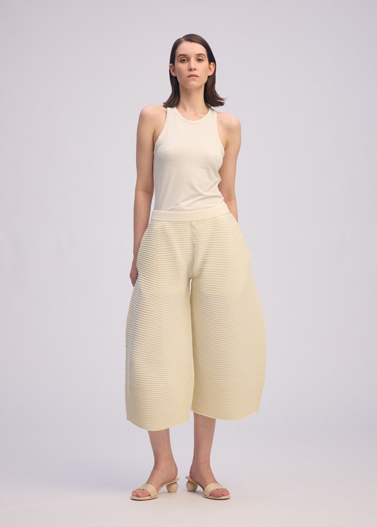 A woman with straight brown hair and a neutral expression stands against a plain background, wearing the ISSEY MIYAKE CORRUGATED KNIT sleeveless beige top, textured wide-leg cream pants, and beige open-toe sandals. view 6 of 8