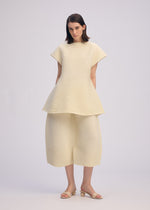 A woman poses against a plain light background wearing the ISSEY MIYAKE CORRUGATED KNIT textured cream short-sleeve top with a flared hem and matching Japanese Washi paper wide-leg pants, paired with beige sandals.