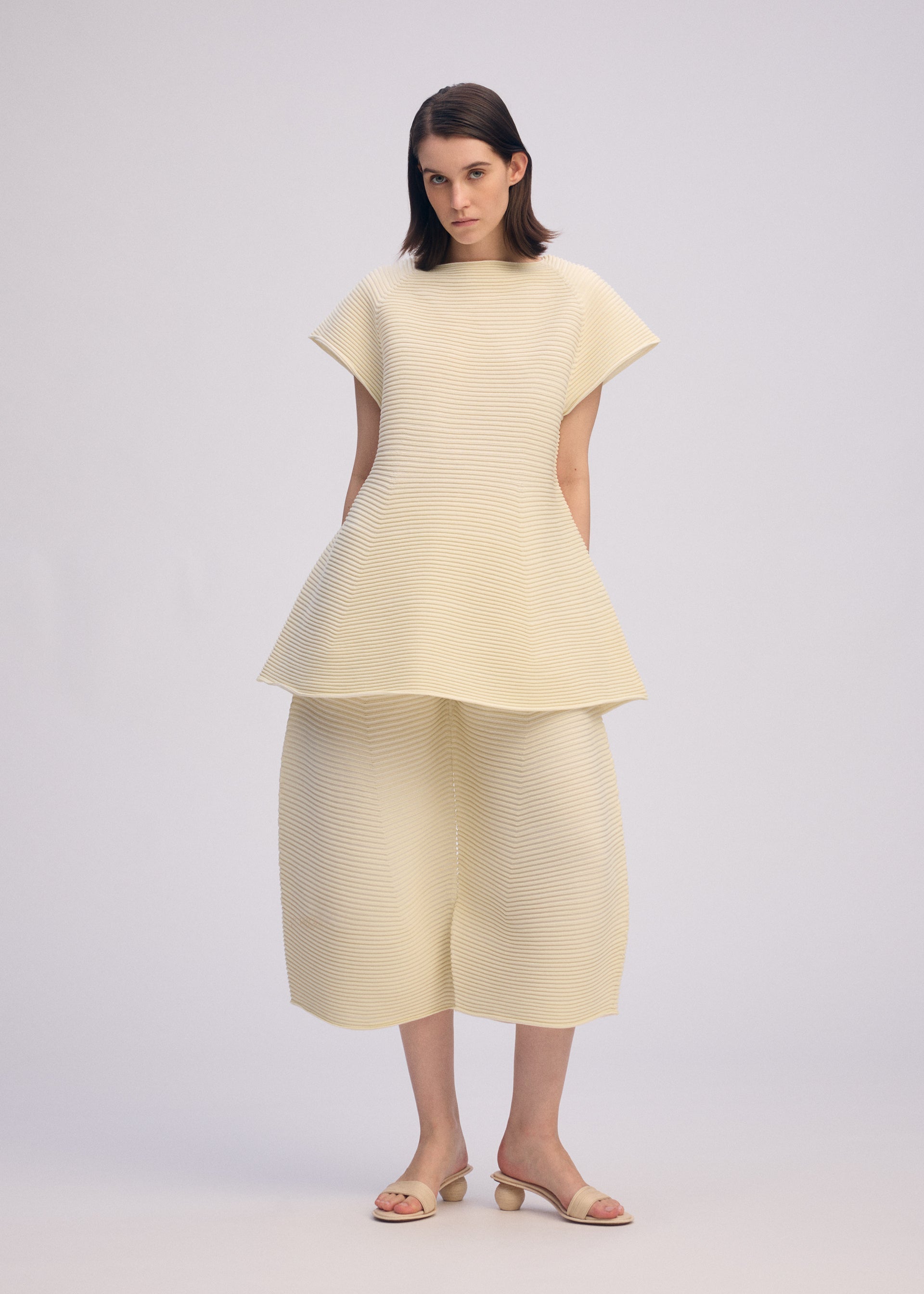 Zoomed view A woman poses against a plain light background wearing the ISSEY MIYAKE CORRUGATED KNIT textured cream short-sleeve top with a flared hem and matching Japanese Washi paper wide-leg pants, paired with beige sandals.