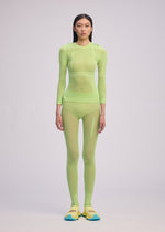 A person stands against a plain background, wearing the ISSEY MIYAKE SLITS MESH AP—a sheer, neon green long-sleeve top paired with mesh knit pants—styled with yellow and blue shoes. Their hair is straight and loose, expression neutral.