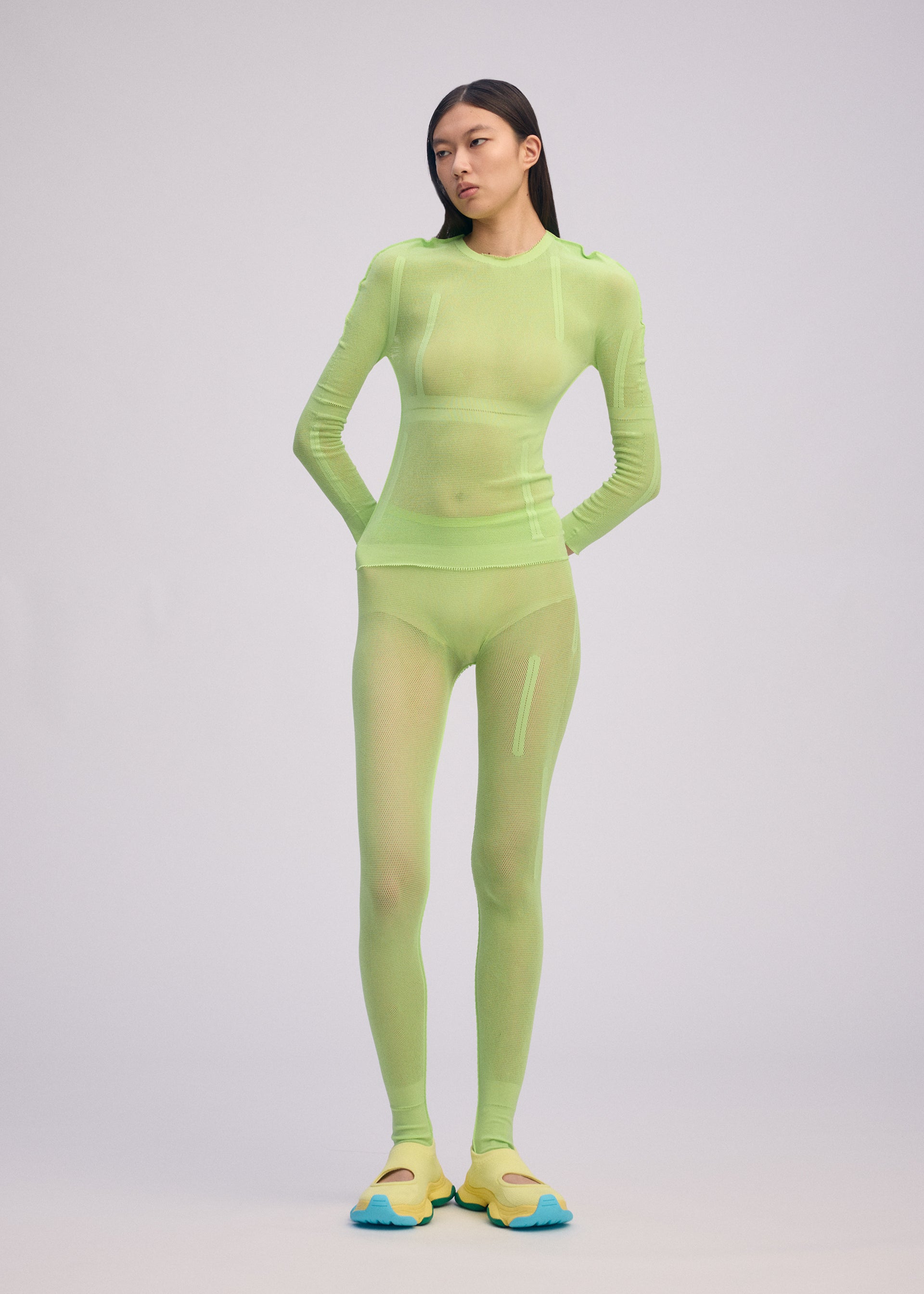 Zoomed view A person with long dark hair and a neutral expression wears the ISSEY MIYAKE SLITS MESH AP—a fitted, sheer neon green long-sleeve top styled with mesh knit pants and yellow-blue shoes against a light grey background. Perfect for layered styling.