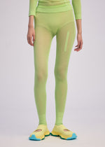 A person wears the ISSEY MIYAKE SLITS MESH AP neon green long-sleeve top and matching mesh knit pants with a vertical pocket, paired with yellow shoes featuring blue soles, standing against a plain light background.