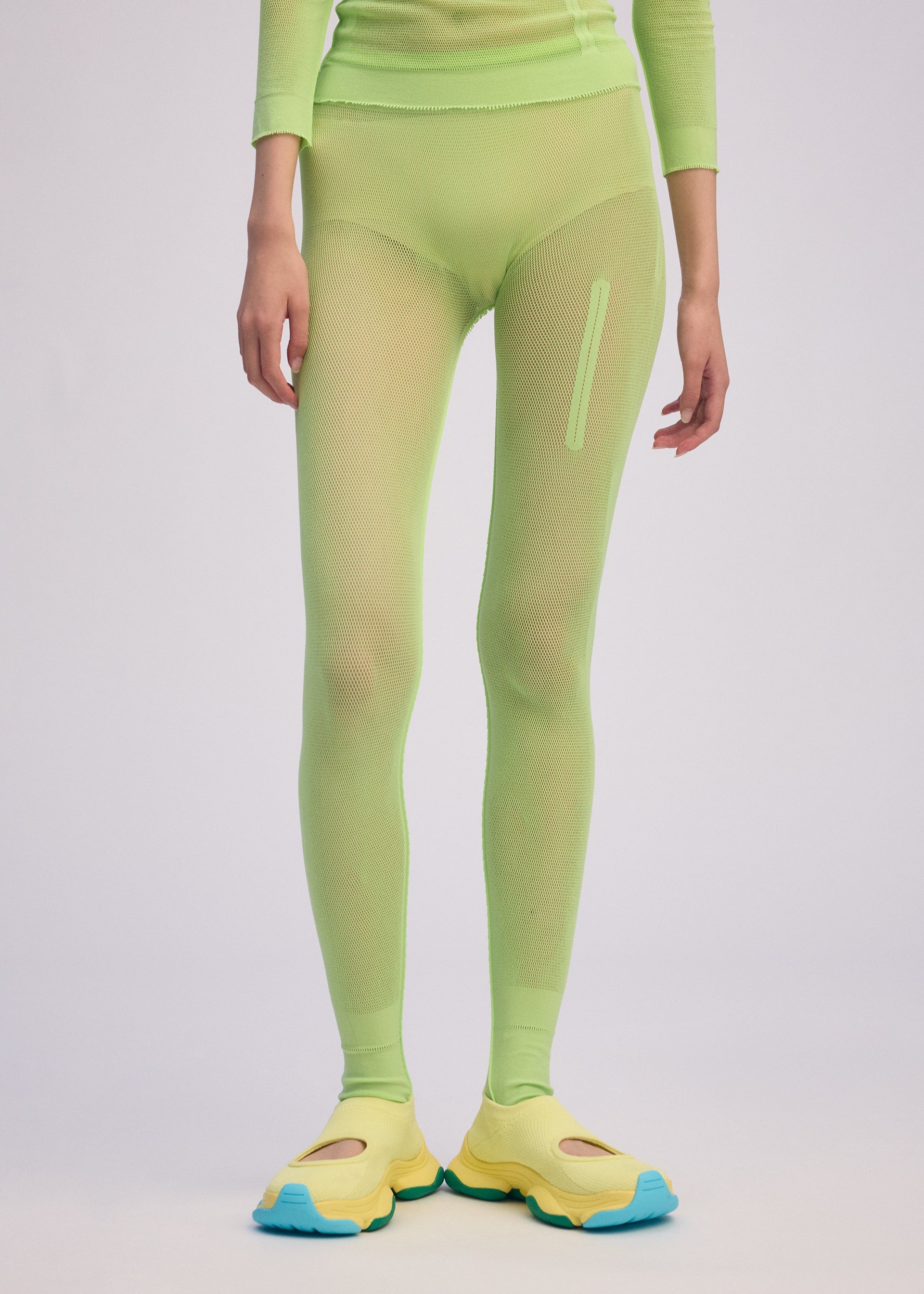 Zoomed view A person wears the ISSEY MIYAKE SLITS MESH AP neon green long-sleeve top and matching mesh knit pants with a vertical pocket, paired with yellow shoes featuring blue soles, standing against a plain light background.