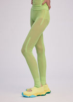 A person stands with one hand on their hip, wearing ISSEY MIYAKE SLITS MESH AP neon green knit pants and yellow slip-on sneakers with blue soles against a plain light background.